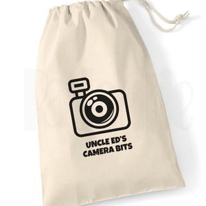 May include: A cream-coloured drawstring bag featuring a black camera graphic and the text "UNCLE ED'S CAMERA BITS". The bag is made of a soft, natural fabric and has a white drawstring closure.