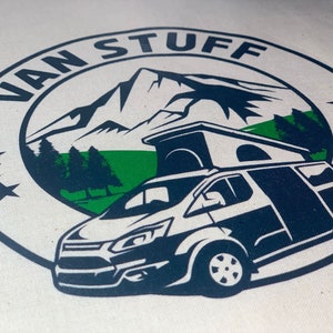 May include: A white canvas tote bag with a blue and green logo. The logo features a mountain range, trees, a camper van, and the text "VAN STUFF".