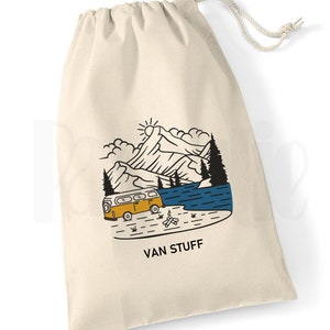 May include: A white canvas drawstring bag featuring a black and yellow illustration of a van driving through a mountain landscape with a lake and a campfire. The text "VAN STUFF" is printed below the illustration.
