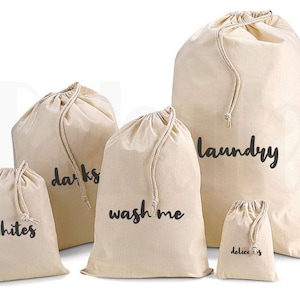 May include: Five beige canvas laundry bags with drawstring closures. The bags are labelled "whites", "darks", "wash me", "delicates", and "laundry" in black script. The bags vary in size, suitable for organising laundry.