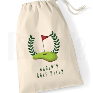 May include: A white canvas drawstring bag with a green golf course and red flag design. The text "Roger's Golf Balls" is printed on the bag.