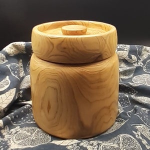 Puede incluir: Un tarro de madera con tapa, hecho de madera natural con un patrón de veta. El tarro es de color marrón claro y tiene una forma redonda y lisa.