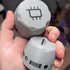 May include: Two gray, cylindrical objects with black graphic designs are held in a hand. The top object has a circuit board symbol, while the bottom one features a series of lines and shapes. The objects appear to be 3D-printed.