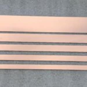 May include: A collection of copper strips in different sizes, displayed on a grey surface. The strips have a polished, metallic appearance and a warm, reddish-brown hue. The longest strip is around 30 cm, and the shortest is about 15 cm in length.