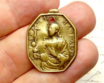 以下が含まれることがあります： 八角形のアンティークゴールドカラーの宗教ペンダント。ペンダントには、十字架とオブジェクトを持つ聖人のエンボス加工された画像があり、周囲には「MA MAGDALP」と「S.S. MA」の文字が刻まれています。