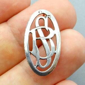 May include: A silver-colored oval pendant featuring an openwork design of intertwined letters, possibly initials. The pendant has a delicate, vintage aesthetic, with a small loop at the top for a chain. The design is set against a neutral background.
