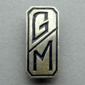 Peut inclure: Une épingle rectangulaire en métal argenté avec les lettres "GM" en noir, dans une police stylisée. Les lettres sont divisées par une ligne diagonale. L'épingle a un bord en relief et un aspect légèrement vieilli.