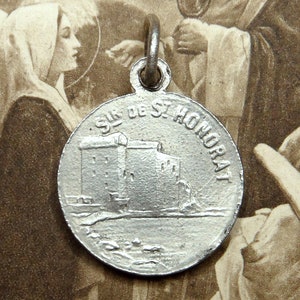 Könnte beinhalten: Ein silberfarbener runder Anhänger mit der Aufschrift "SUR DE ST. HONORAT". Der Anhänger zeigt eine detaillierte Darstellung eines Gebäudes mit Türmen und einem Gewässer.