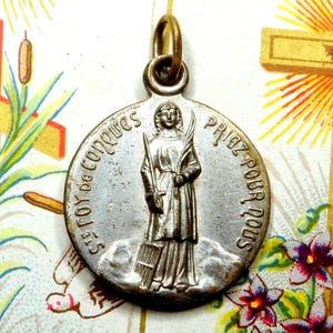 May include: A circular silver-toned metal religious pendant featuring a raised relief of a figure holding a palm frond and a grid-like object. The inscription reads St. Foy de Conques Priez pour nous. A small metal jump ring is attached at the top.