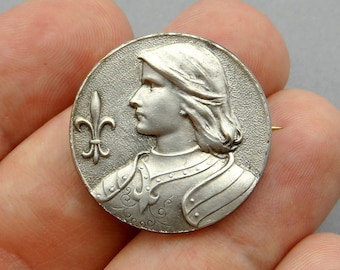 May include: Silver tone metal pin featuring a relief sculpture of a knight in armor with a fleur-de-lis. The knight is facing left and has a long, flowing mane of hair.