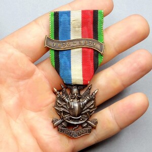 Puede incluir: Una medalla vintage con una cinta colorida con rayas verdes, negras, azules, blancas y rojas. La medalla tiene un emblema de color bronce con espadas cruzadas y el texto "1870-1871" y "JOUER... JAMAIS!"
