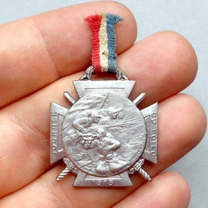 May include: A silver medal with a cross design and a depiction of soldiers in combat. The medal reads "JOURNEE DU POILU 1915".