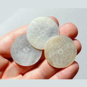 Peut inclure: Trois pièces décoratives rondes, blanc cassé, avec des motifs floraux. Chaque pièce présente une fleur gravée avec des volutes. Les pièces sont tenues dans une main, mettant en valeur leurs détails délicats et leurs subtiles variations de couleur.