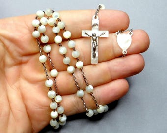 May include: A rosary with white, round beads and a silver chain. The rosary includes a white cross with a silver Jesus figure and a white heart-shaped pendant.