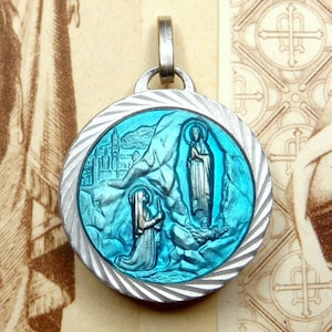 Puede incluir: Pendiente de plata con una imagen de esmalte azul de la Virgen María apareciéndose a una mujer arrodillada en una cueva. El pendiente tiene un borde decorativo texturizado.