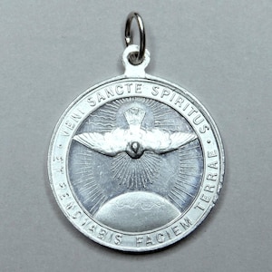 Könnte beinhalten: Runder, silberfarbener Anhänger mit einer Taube und der Aufschrift "VENI SANCTE SPIRITUS ET RENOVABIS FACIEM TERRAE". Die Taube befindet sich über einem Sonnenstrahl-Design. Oben befindet sich eine kleine Schlaufe für eine Kette.