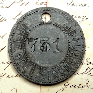 May include: A circular metal tag with a hole at the top. The tag is grey and has the text "GEF. L. STROHER MOOR" and the number "731" printed on it.
