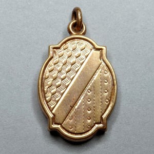 Peut inclure: Un médaillon ovale doré avec un motif décoratif comprenant une ligne diagonale et un motif ondulé. Le médaillon a une boucle en haut pour attacher une chaîne.