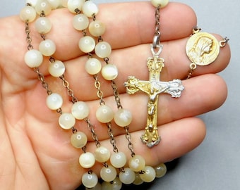 May include: A vintage rosary with luminous, round, off-white beads strung on a delicate chain. The rosary features a gold-toned crucifix with a silver figure of Jesus and a gold-toned medallion with a profile of a religious figure.