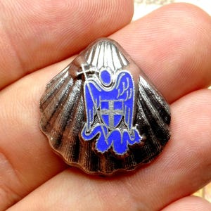Peut inclure: Une épingle en forme de coquillage argenté avec un emblème bleu et argenté. L'emblème représente un ange stylisé avec une croix. L'épingle est probablement en métal et a une surface texturée et détaillée.