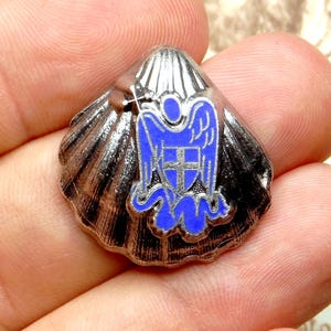 Peut inclure: Une épingle en forme de coquillage argenté avec un emblème en émail bleu représentant un ange et une croix. L'épingle a une texture détaillée et striée et est tenue dans une main.
