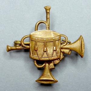 Op de afbeelding: Een goudkleurige metalen badge met een trommel in het midden, geflankeerd door trompetten en een hoorn. De trommel heeft ingewikkelde details en de badge heeft een vintage uitstraling. Het ontwerp suggereert een militair of bandthema.