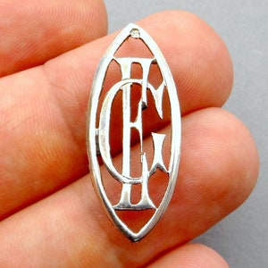 May include: A silver pendant with a cutout design of the letters "CE" in a stylized font. The pendant is oval shaped and has a smooth, polished finish.