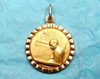 May include: Gold pendant with a detailed image of a person praying in front of a cross. The pendant is round and has a textured edge.