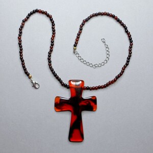 Peut inclure: Un collier avec un grand pendentif en forme de croix rouge et marron. Le collier est composé de perles brunes et noires.