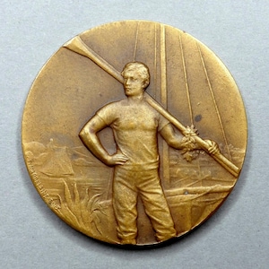 May include: A bronze-colored round medal featuring a man holding a long pole over his shoulder. The man is depicted in a t-shirt and pants, with one hand on his hip. The background shows a landscape with structures and vegetation.