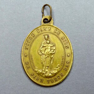 Peut inclure: Un pendentif en métal doré avec une image religieuse de la Vierge Marie et de l'enfant Jésus. Le pendentif est inscrit avec le texte "NOTRE DAME DE SION DE SION TUBLIE BAS".