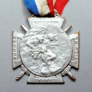 May include: A silver medal with a cross design and a circular center depicting a scene of soldiers in action. The medal is inscribed "JOURNEE DU POILU 1915".