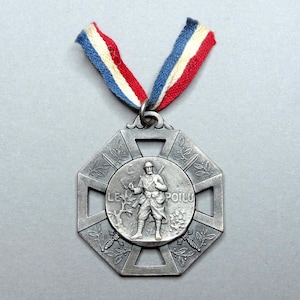 May include: A silver-toned octagonal medal featuring a central relief of a soldier with the text LE POILU. The medal is suspended from a fabric ribbon with vertical stripes in blue, white, and red. The design includes engraved laurel branches.