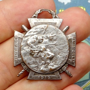 May include: A silver metal cross-shaped medal with the words "JOURNEE DU POILU 1915" inscribed on it. The medal features a raised image of soldiers in a battle scene.