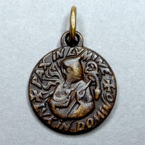 May include: A bronze-toned pendant with a circular design. The pendant features an embossed image of a figure holding an object, surrounded by text. The pendant has a small loop at the top for hanging.
