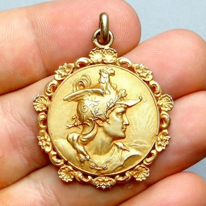 May include: A gold-colored pendant featuring a detailed relief of a classical figure in profile, wearing a helmet adorned with a rooster. The pendant is circular, with a floral border and a loop for a chain. A vintage jewelry piece.