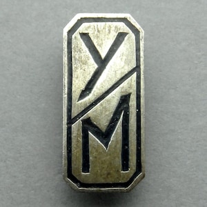 Peut inclure: Une épingle en métal argenté avec les lettres "YM" dans une police de caractères stylisée. Les lettres sont noires et l'arrière-plan est argenté.