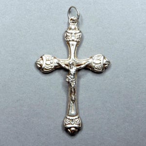 Può includere: Ciondolo a forma di croce color argento con una dettagliata raffigurazione di Gesù Cristo. La croce presenta dettagli decorativi sulle braccia e sulla base, e un piccolo anello per una catena. Il ciondolo è esposto su sfondo grigio.