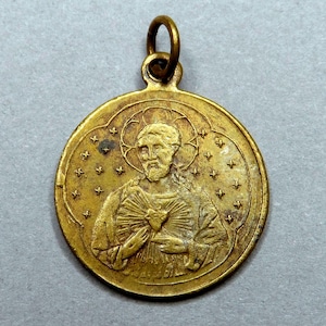 Puede incluir: Colgante dorado con una imagen detallada de un hombre con barba, sosteniendo su mano sobre su pecho. El colgante tiene un halo y está rodeado de pequeños diseños en forma de estrella. Un pequeño lazo en la parte superior para una cadena.