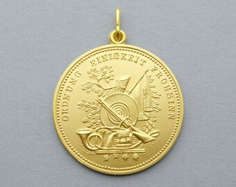 May include: A gold-colored pendant with a circular design. The pendant features embossed text and detailed imagery of musical instruments, flags, and foliage. The words "EINIGKEIT FROHSINN" and "ORDNUNG" are visible on the pendant.