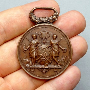 Puede incluir: Una medalla de bronce con un lazo en la parte superior. El anverso de la medalla presenta a dos mujeres de pie una al lado de la otra, cada una sosteniendo una corona de laurel. Las mujeres están de pie sobre una plataforma con un escudo y otros símbolos.