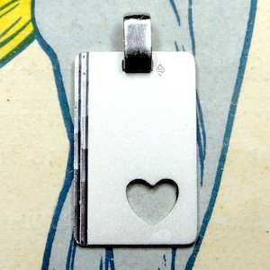 Peut inclure: Pendentif argenté en forme de petit livre avec une découpe en forme de cœur. Le pendentif a une forme rectangulaire avec une boucle pour une chaîne. Les bords du livre sont détaillés.