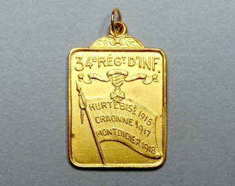 May include: A rectangular gold-colored pendant with a loop at the top. The pendant features the text "34e REGT D'INF." and a banner with the text "HURTEBISE 1915, CRAONNE 1917, MONTDIDIER 1918".