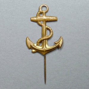 Puede incluir: Un pin de ancla de metal de color dorado con un diseño detallado. El ancla tiene un detalle de cuerda en la parte superior y una punta afilada en la parte inferior.
