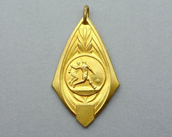 May include: A gold-colored medal with a design of two figures playing soccer. The medal is in the shape of a teardrop with a pointed bottom.