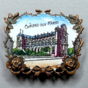 Op de afbeelding: Een vintage porseleinen broche met een gedetailleerd beeld van een gebouw en de tekst "Châlons-sur-Marne". De broche is omgeven door een decoratief metalen frame met een rozenontwerp.