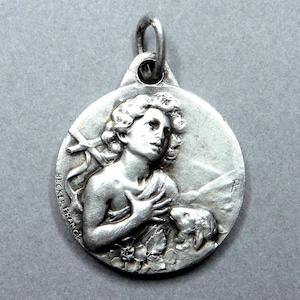 Op de afbeelding: Een ronde zilverkleurige metalen hanger met een reliëf van een figuur met krullend haar, één hand op de borst en een klein dier ernaast. De tekst BECKER FRANCE staat in reliëf aan de linkerkant van de ronde medaille.