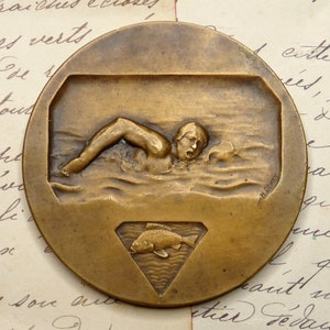 May include: Bronze medal featuring a swimmer in a stylized depiction of water. The medal also has a fish and a diamond shape in the lower portion. The medal is signed "H. Dizery".