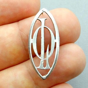Peut inclure: Pendentif argenté de forme ovale allongée. Le pendentif présente un motif ajouré avec les lettres "IOH" entrelacées. Les lettres sont dans une police stylisée. Le pendentif est tenu entre les doigts. Le métal est brillant.