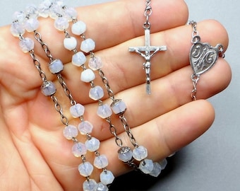 May include: A rosary with light-colored, faceted beads and silver-toned chain links. The rosary features a crucifix and a heart-shaped pendant with an ornate design. The beads appear to be made of a translucent material.